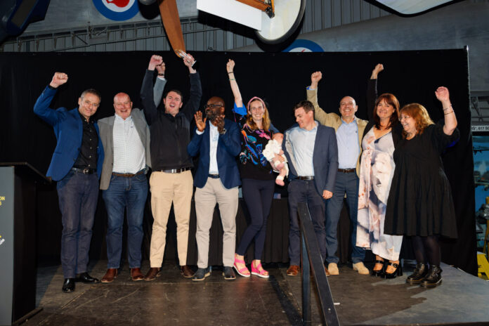 A group of people are standing on a stage. They are all raising their hands up in the air. There is a woman in a black dress standing next to them.